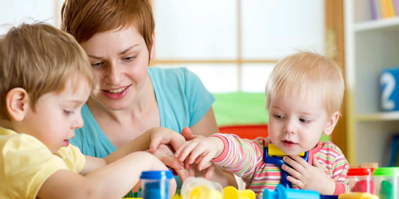 Kita-Helferin spielt mit zwei Kleinkindern am Tisch